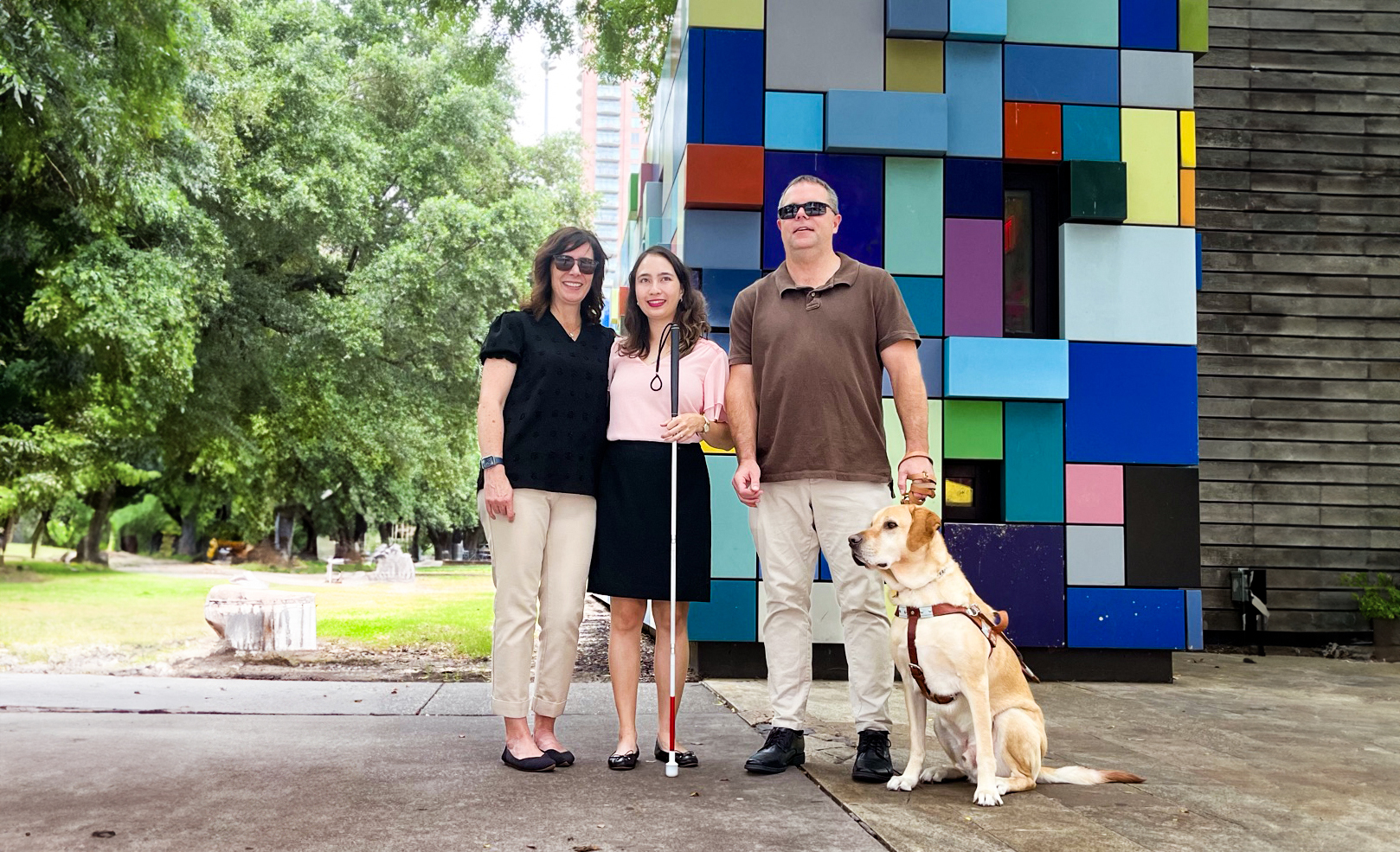 Three people standing outside in front of a piece of colorful art; the person on the far left is wearing sunglasses, the middle person is holding a cane, and the person on the right is also wearing sunglasses and is holding the leash of his dog guide.