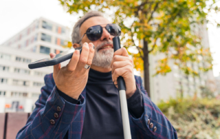 A man with greying hair is standing outside, wearing sunglasses. In one hand he is holding a white cane and in the other a smartphone.