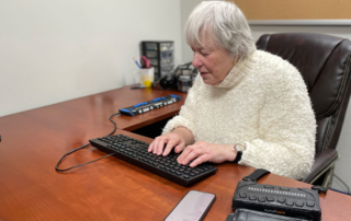 A woman with light skin and white hair sits at a desk, typing on a keyboard.