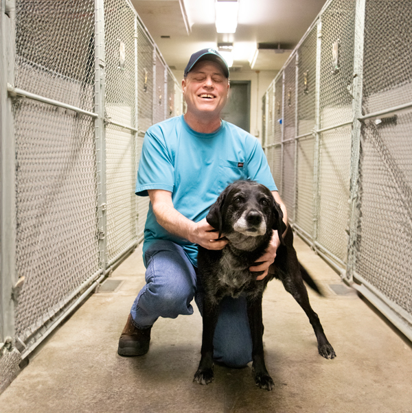 Mike, a light-skinned man wearing a baseball cap smiles and embraces his dog guide in a kennel at the Seattle facility