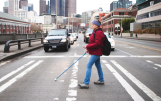 A woman crossing the street using a white cane and a mini-guide.