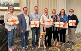 7 people stand holding certificates. One person is holding a white cane and another has a dog guide.