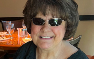 Photo of Julie, a light-skinned woman wearing dark sunglasses sitting inside of a restaurant and smiling