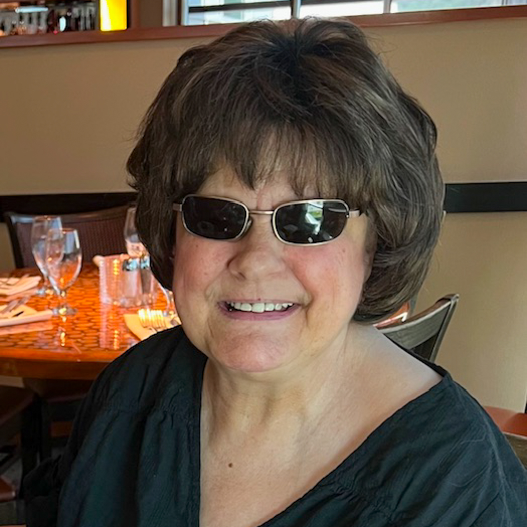 Photo of Julie, a light-skinned woman wearing dark sunglasses sitting inside of a restaurant and smiling
