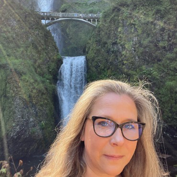 Photo of Jen Smith, a light-skinned woman with glasses, standing outside in front of a waterfall