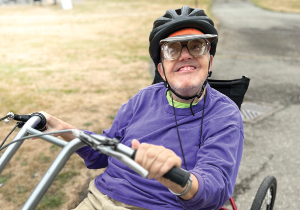 Scott Oberg, DeafBlind Retreat camper. Scott has light skin and is smiling while sitting on a low tricycle, wearing a bike helmet.