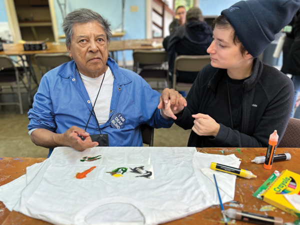 Roy Rios, DeafBlind Retreat camper, and Sydney Tidler, volunteer Interpreter. Roy has medium skin and a grey hair and is sitting at a craft table with a t-shirt and markers in front of him.