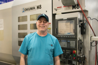 Mike King, CNC Machinist II, Seattle Facility. Mike has light skin and it standing in front of a large AKUMA machine, wearing a baseball cap and smiling.