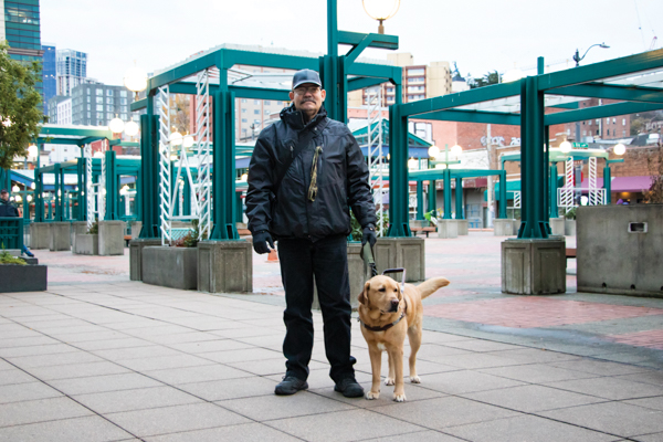 John Jeans, Maching Operator, Seattle Facility. John has light skin and a large brown mustache. He is standing outside on a sidewalk next to his dog guide.