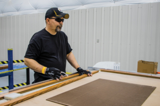 John Jack Lloyd, Production Worker, Spokane facility. John has medium skin, is wearing a hat, dark sunglassed, and protective gloves as he works at an assembly table.