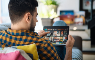 A man sits on a couch, scrolling through a streaming service on his tablet.