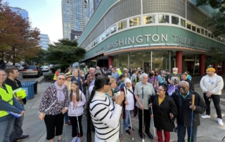 A large group of people stand in front of the WA Talking Book & Braille Library with white canes.