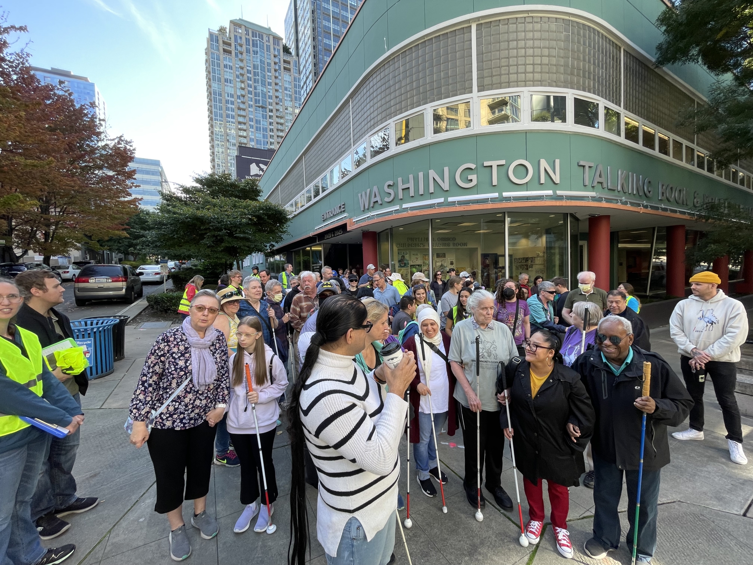 A large group of people stand in front of the WA Talking Book & Braille Library with white canes.
