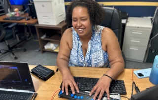 A black woman sits at desk typing on a keyboard braille display, smiling.