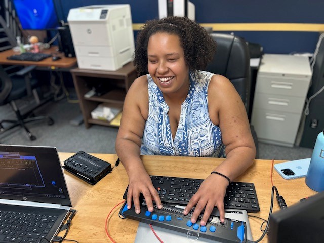 A black woman sits at desk typing on a keyboard braille display, smiling.