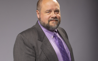 Light-skinned man stands smiling at the camera, wearing a suit and tie.