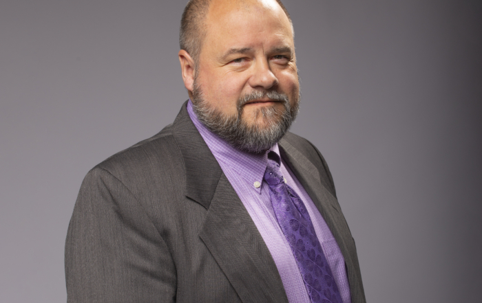 Light-skinned man stands smiling at the camera, wearing a suit and tie.
