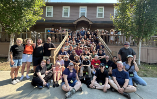 a group of nearly 100 DeafBind Retreat Campers sit on stairs in front of a lodge.