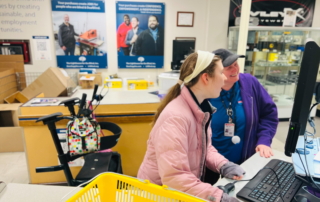 Two women stand looking at a computer screen, scanning in items, with a shopping basket in the foreground.