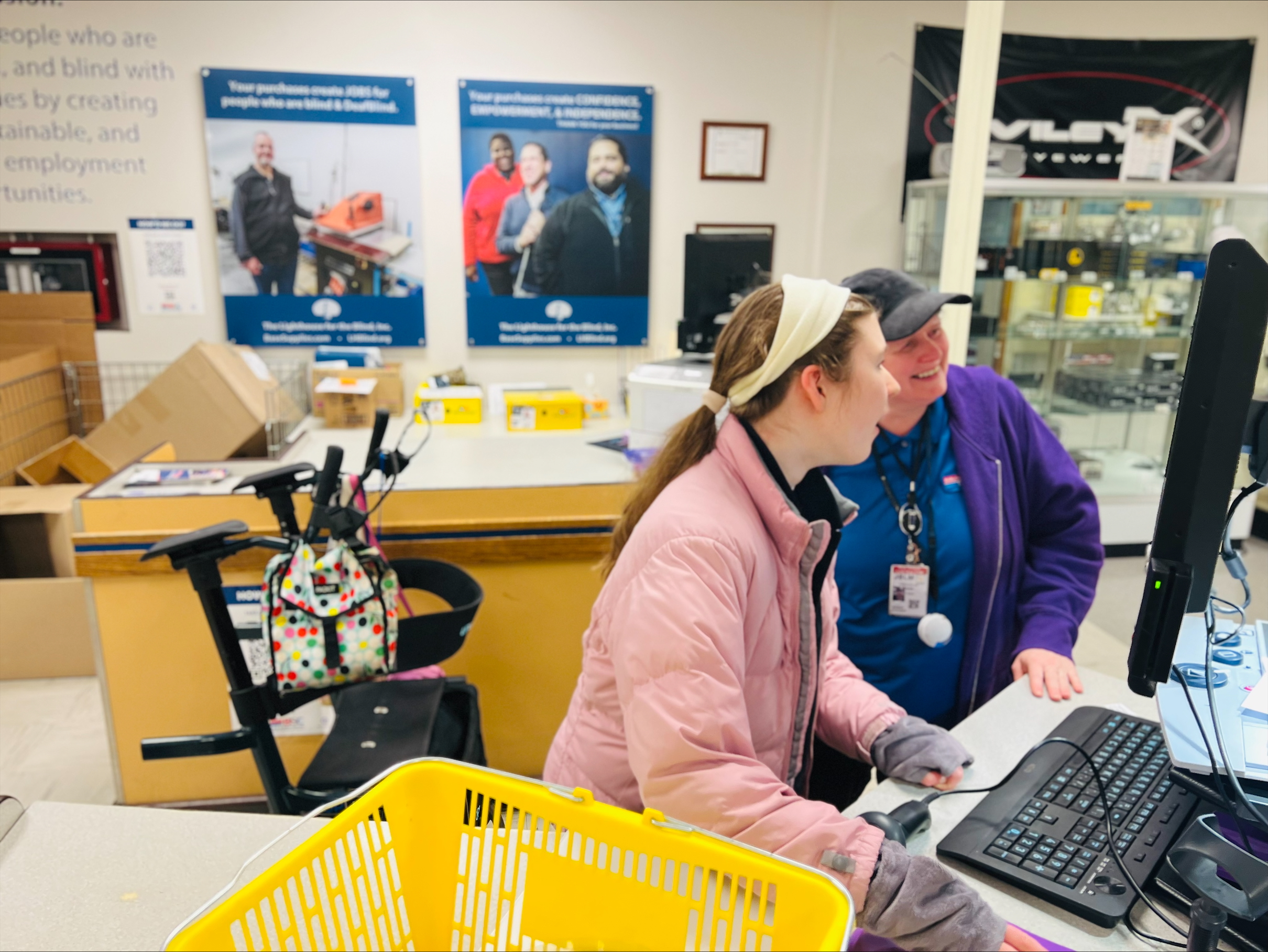 Two women stand looking at a computer screen, scanning in items, with a shopping basket in the foreground.