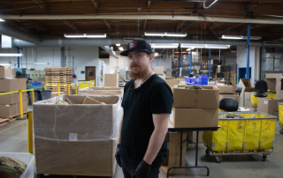 Gregory Wilson standing on the Manufacturing floor, with boxes all around him.