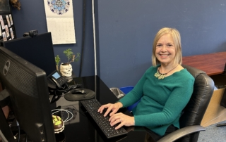 A woman with blonde hair and a green sweater sits at a workstation and smiles.