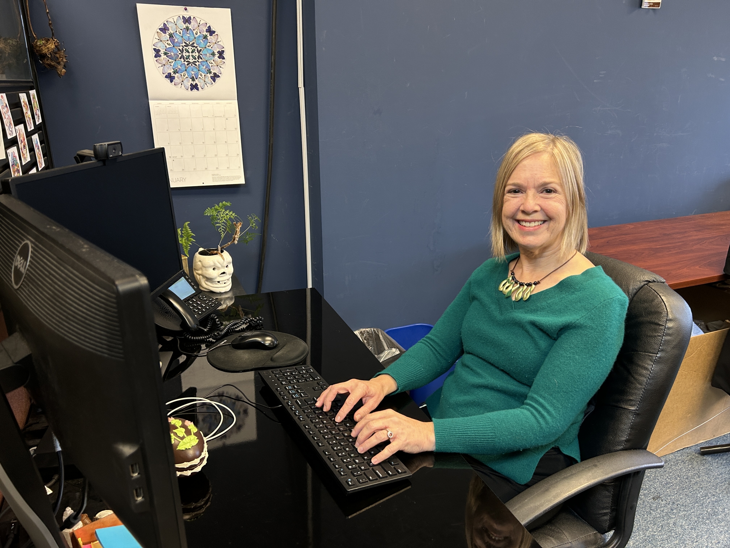 A woman with blonde hair and a green sweater sits at a workstation and smiles.