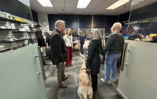a group of people stand in the low vision clinic and store talking. One person is holding a cane, another has a dog guide.