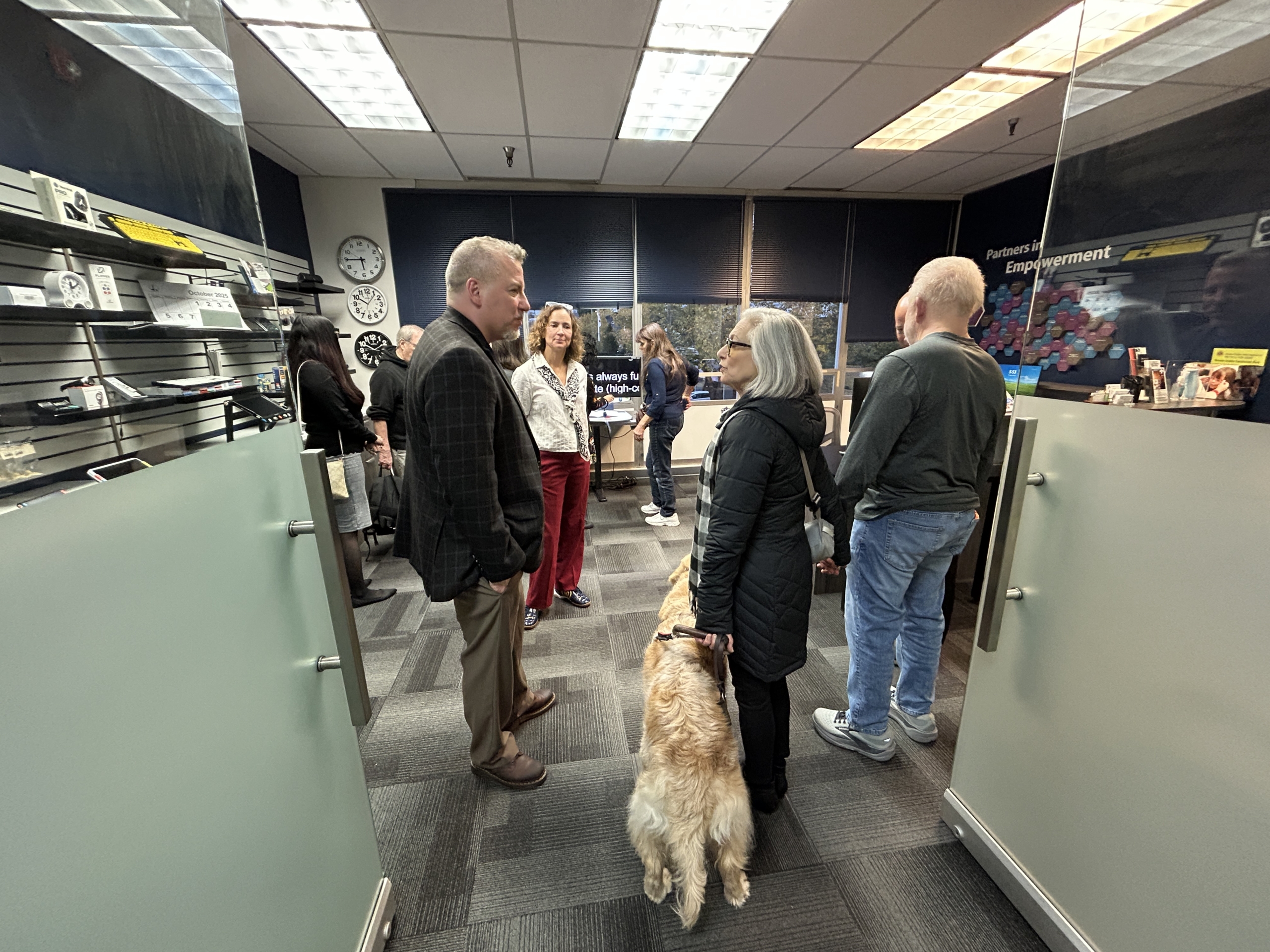 a group of people stand in the low vision clinic and store talking. One person is holding a cane, another has a dog guide.