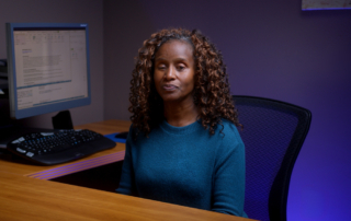 Photo of Cathy, a dark-skinned woman sitting at her computer workstation and smiling