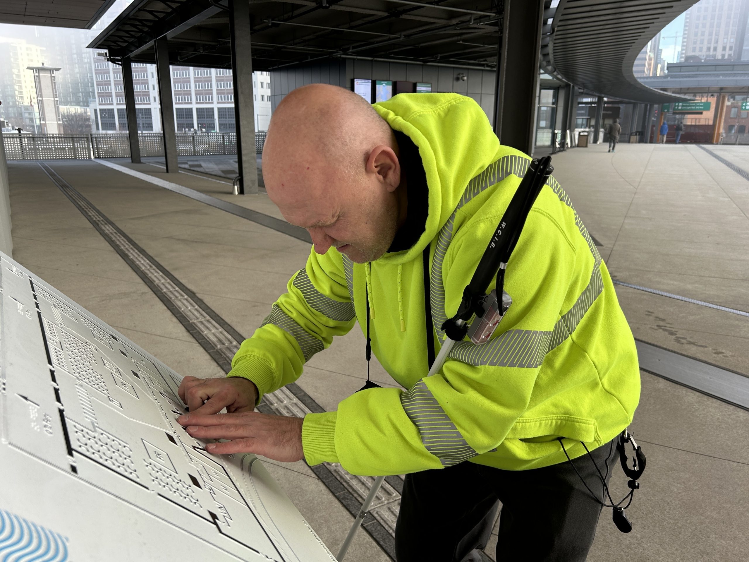 Chris Loomis stands at a kiosk reading Braille in a bright yellow reflector jacket