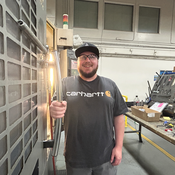 A man wearing a black shirt and safety glasses smiles and holds on to the door of a CNC machine