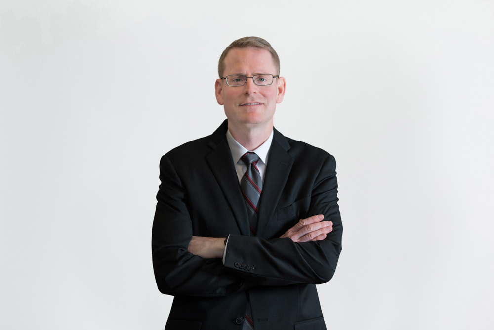 John Craddock, wearing a suit and tie and glasses, smiles in a photo studio