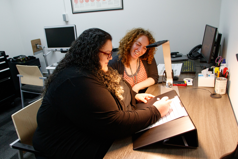 Two women are sitting at a desk and smiling while one is demonstrating accessible technology to the other