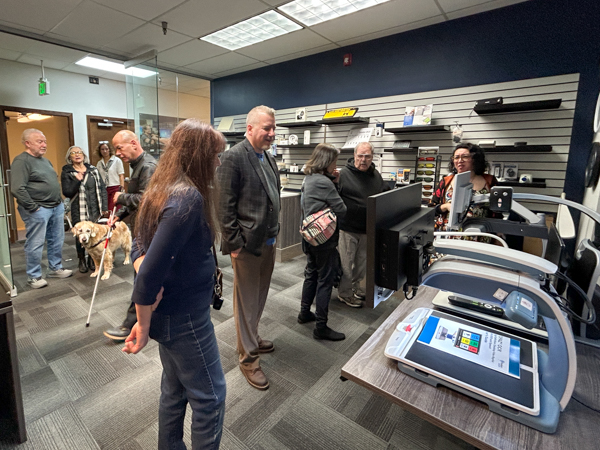 A group of people are standing in a room, looking at a computer screen. There are several chairs and a dog in the room