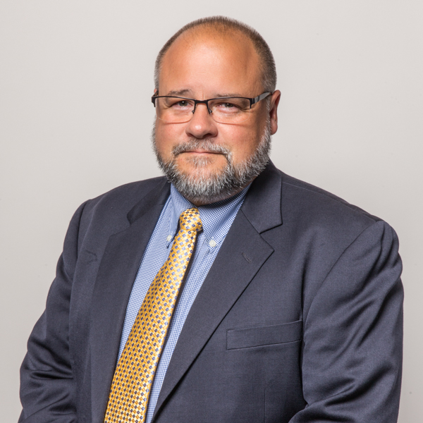 A bearded man wearing glasses and a suit and tie smiling in a photo studio