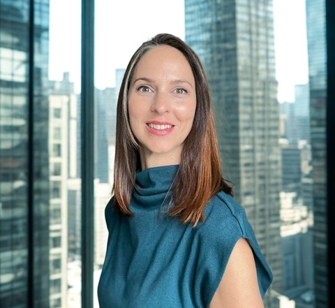 A woman in a teal colored blouse, shoulder length hair, stands in front of a large window with the city in the background
