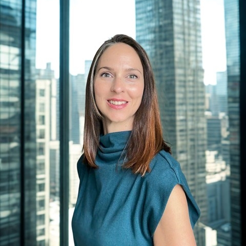 A woman in a teal colored blouse, shoulder length hair, stands in front of a large window with the city in the background