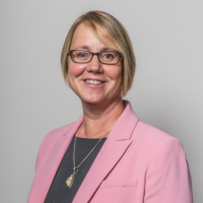 AmyKoehl2019web-1 Amy Koehl standing smiling in a pink suit coat, gray shirt; her hair is blond shoulder length, and is wearing glasses