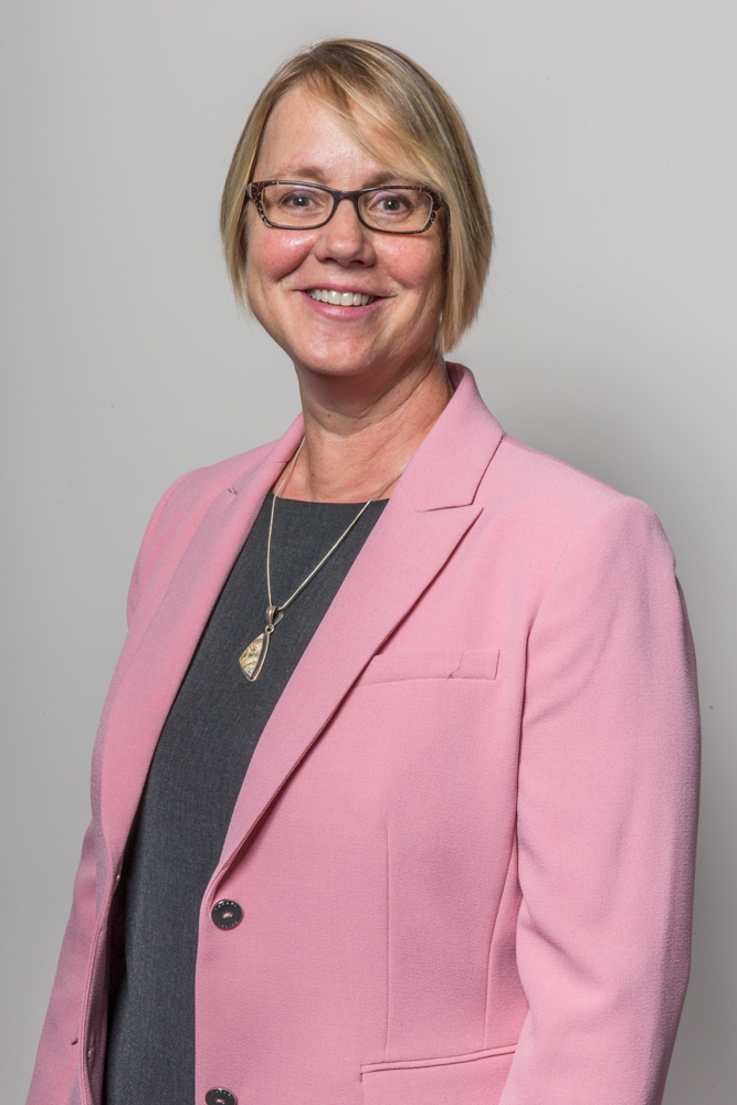 Amy Koehl standing smiling in a pink suit coat, gray shirt; her hair is blond shoulder length, and is wearing glasses